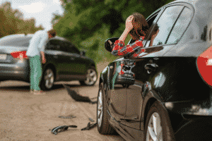 Scène d'accident : Femme dans sa voiture accidentée se tenant la tête ett homme au loin, sur la route, constatant les dégâts sur son véhicule.