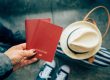 main de femme tenant deux passeports rouge. Un chapeau et un sac par terre en arrière plan